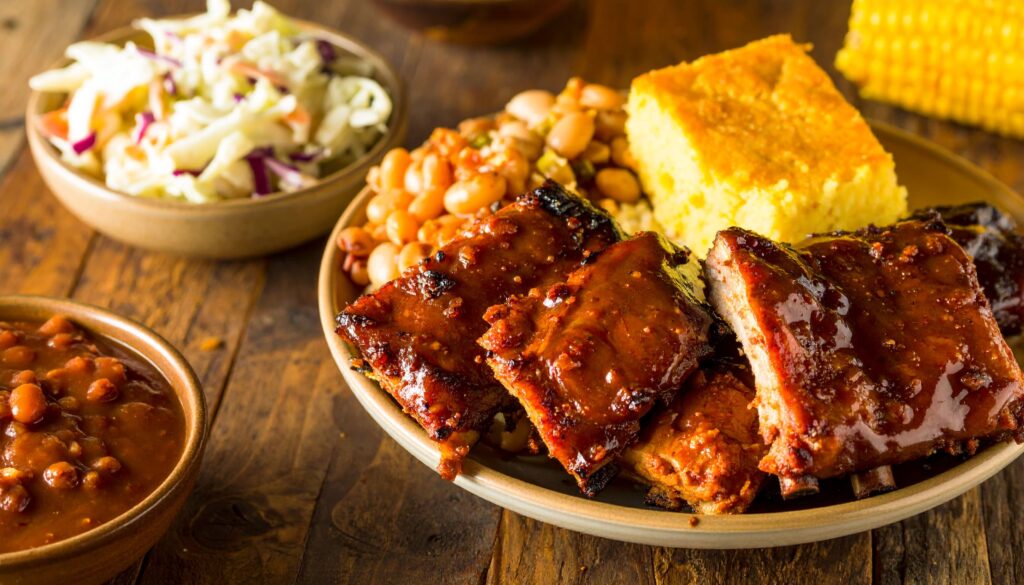 Nahaufnahme von glasierten BBQ-Rippchen mit Beilagen im rustikalen Südstaatenstil, auf Holztisch mit warmem Sonnenlicht.